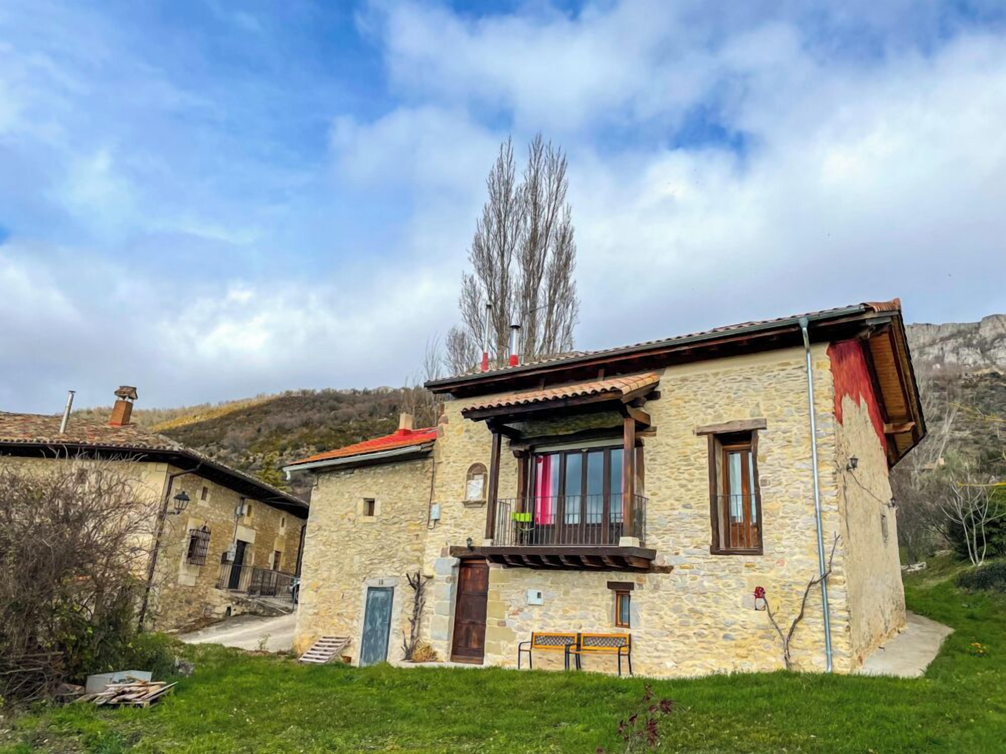 A stone house with a balcony, a small garden, and a mountain backdrop.