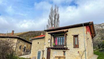A stone house with a balcony, a small garden, and a mountain backdrop.