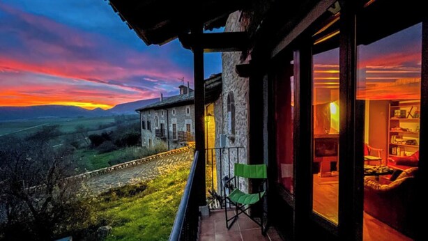 A balcony with a view of a sunset over a rural landscape, a stone building, and a room with a large window. - Casa Berrobiaenea  (Lana)