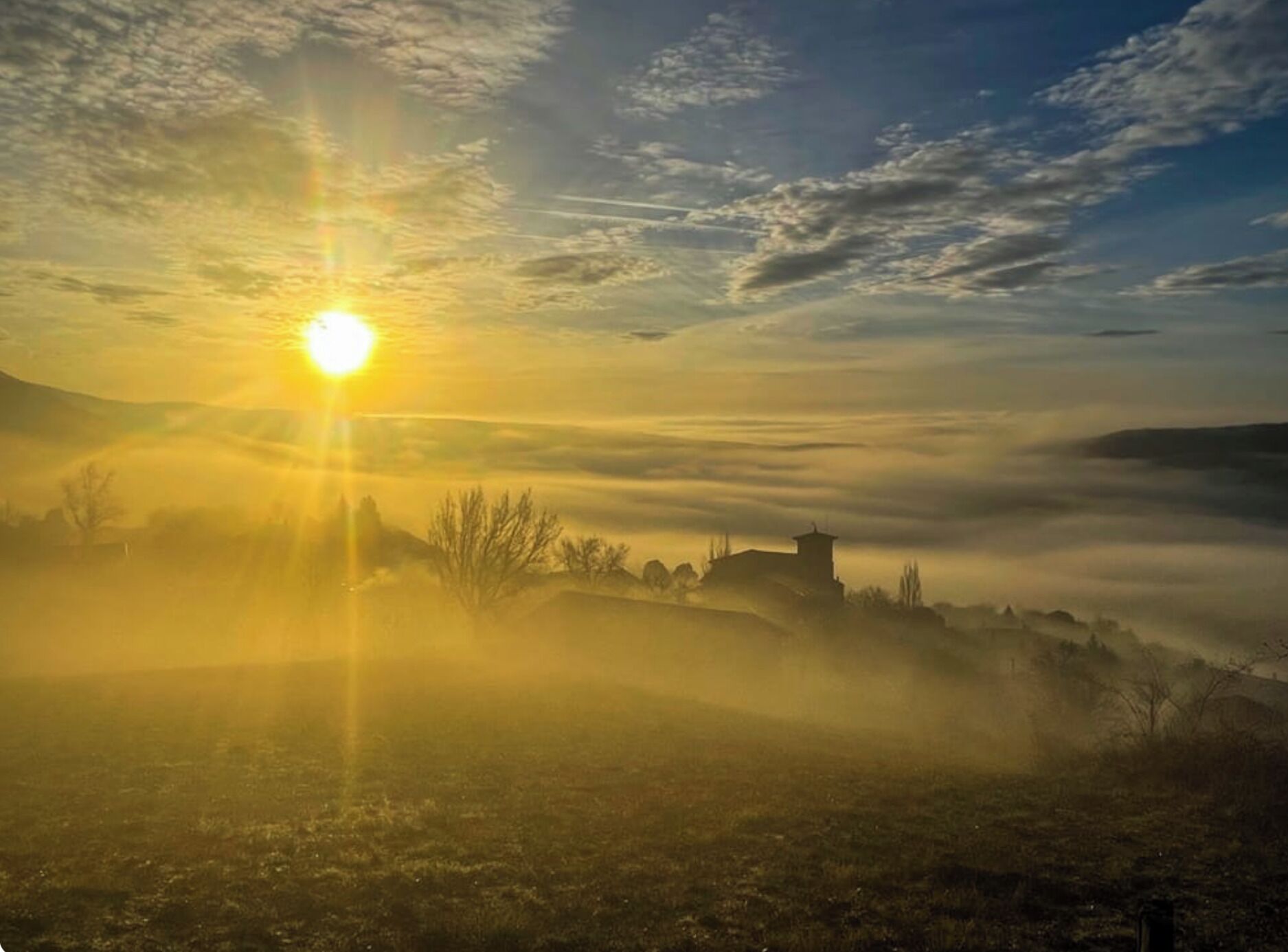 A sunrise over a misty landscape with a solitary building in the distance.