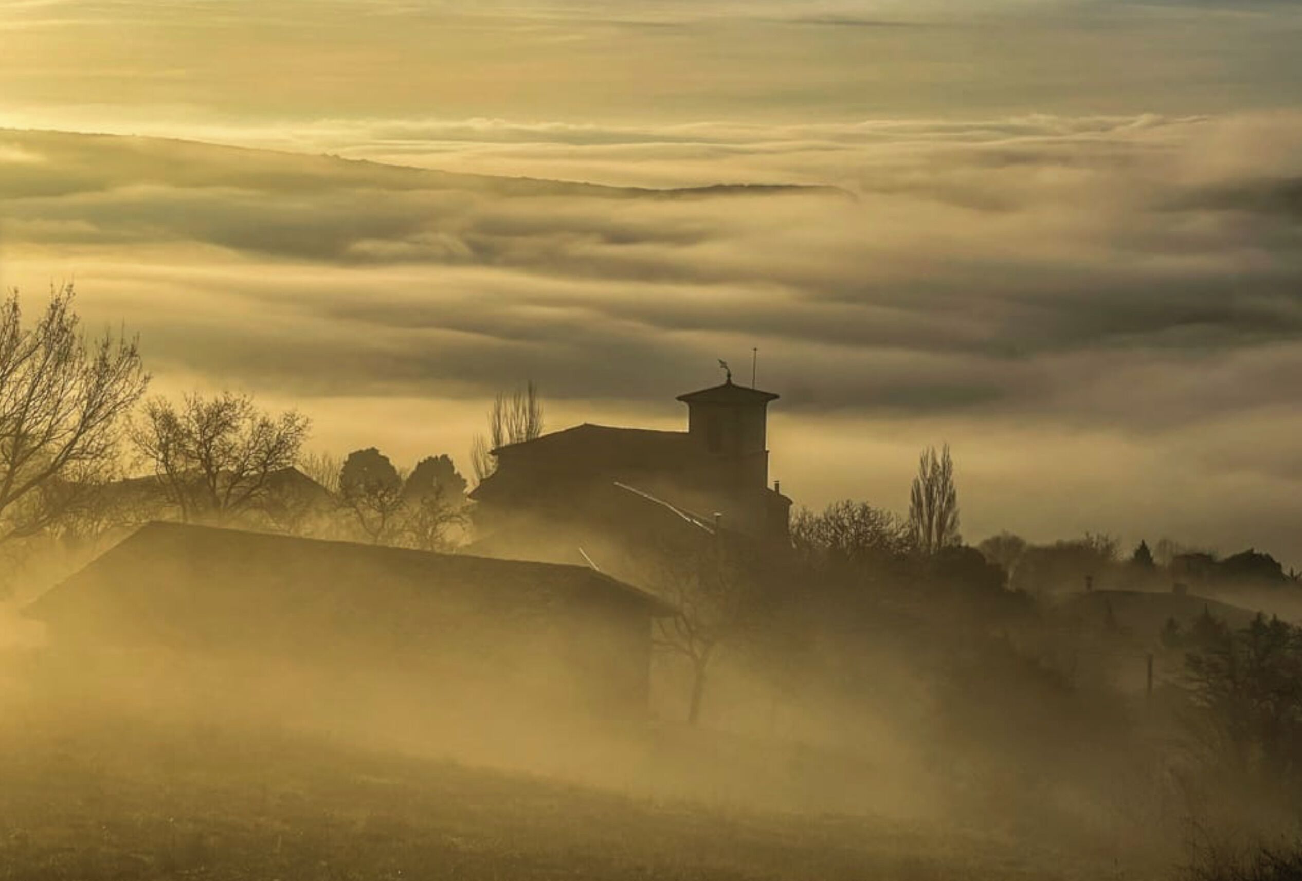 A foggy landscape with a building that has a tower and a flat-roofed structure.