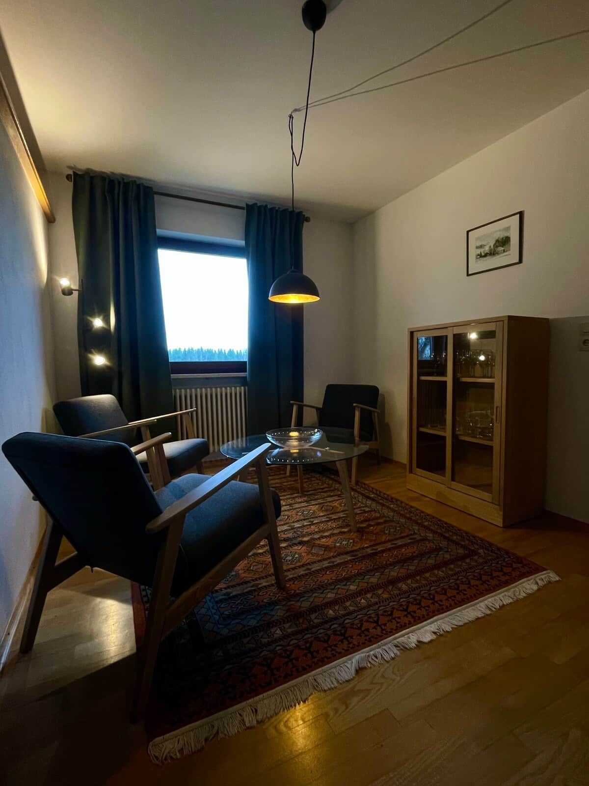 A living room with a dining table, chairs, a hanging light fixture, a cabinet, and a window with curtains.