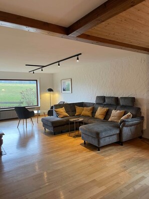 A modern living room with a sectional sofa, a small table, and a chair. There is a large window providing a view of the outdoors.