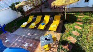 Un espace aménagé au bord de la piscine, avec des chaises longues jaunes et une table, le tout entouré d’un jardin agrémenté de pierres plates servant de chemin et d’un petit arbre.