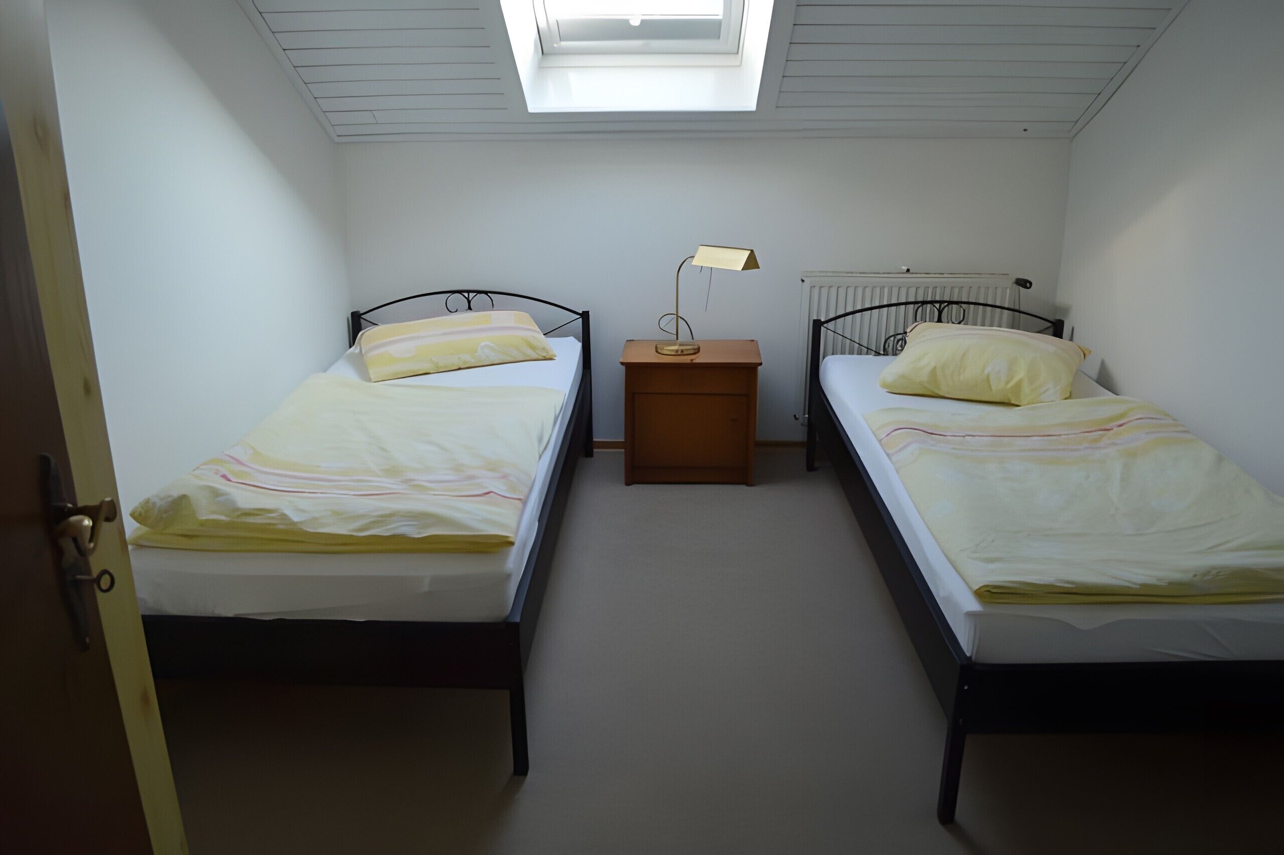 Ein Schlafzimmer mit zwei Einzelbetten, einem Nachttisch mit Lampe, einem Heizkörper und einem Oberlicht.