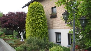 A building with a brick facade, a tall hedge, and a well-maintained garden with various plants and flowers. - Apartment 'Almer' with Balcony and Wi-Fi (Brilon)