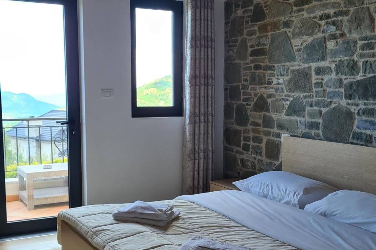A bedroom with a stone wall, a bed with white linens, and a view of a balcony with a table and chairs.