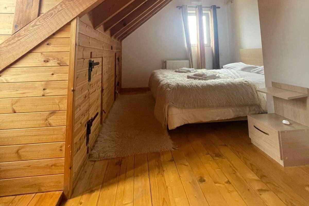 A cozy attic bedroom with wooden beams, a sloped ceiling, a bed with a fluffy blanket, and a small desk with a computer.