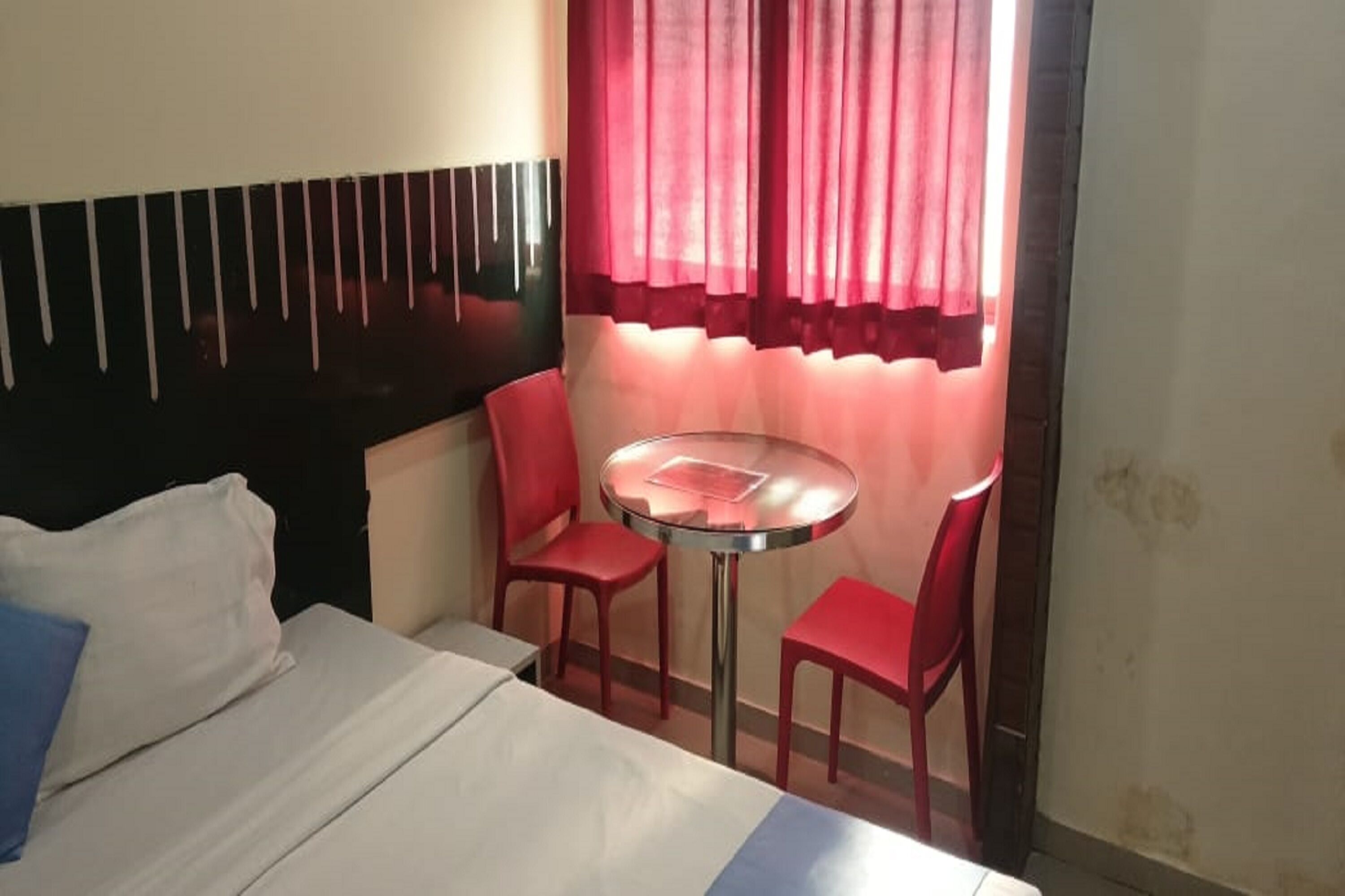 A hotel room with a bed, a small round table, two red chairs, and a window with red curtains.