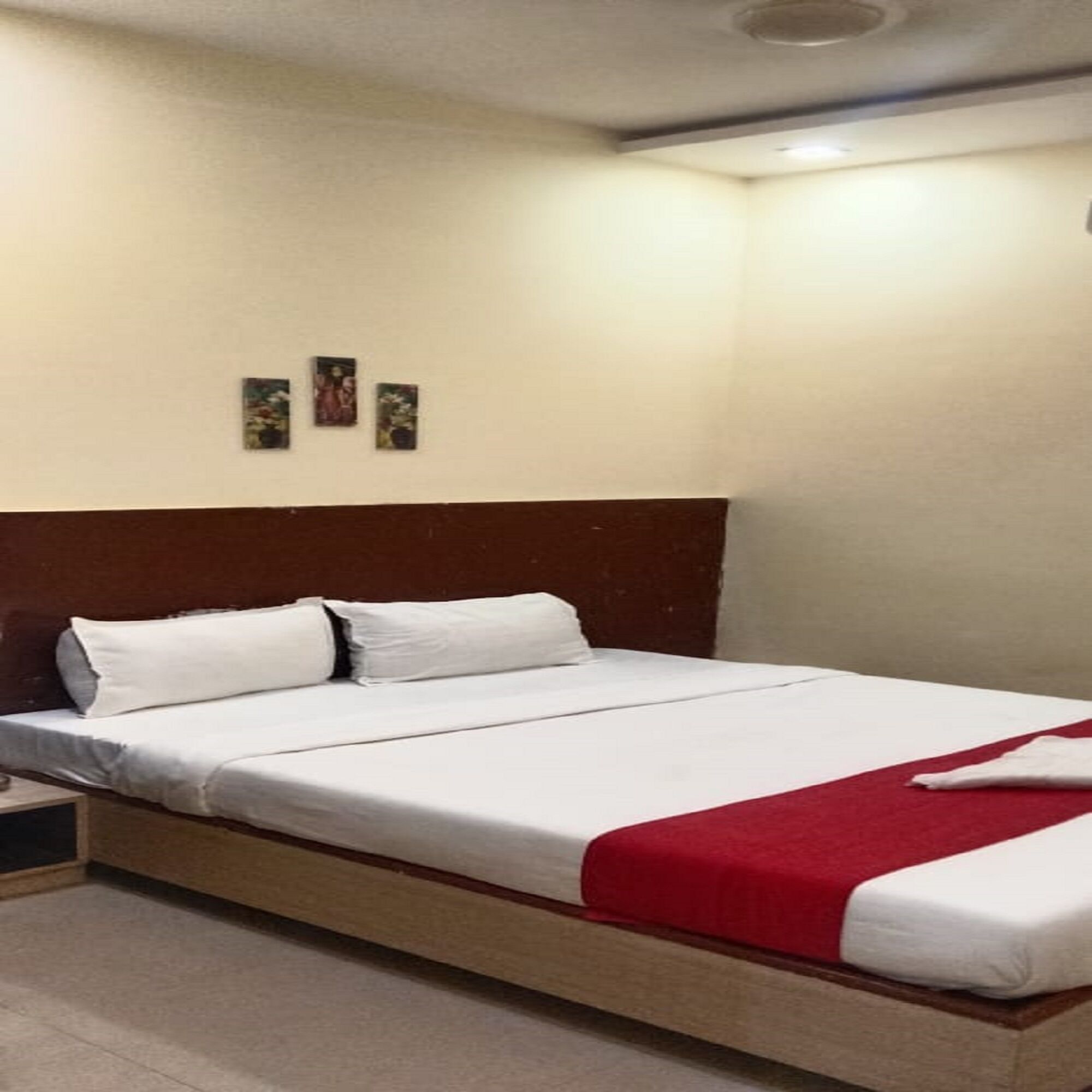 Una cama bien hecha con sábanas blancas y una manta roja, un cabecero de madera y tres pequeños cuadros enmarcados en la pared.