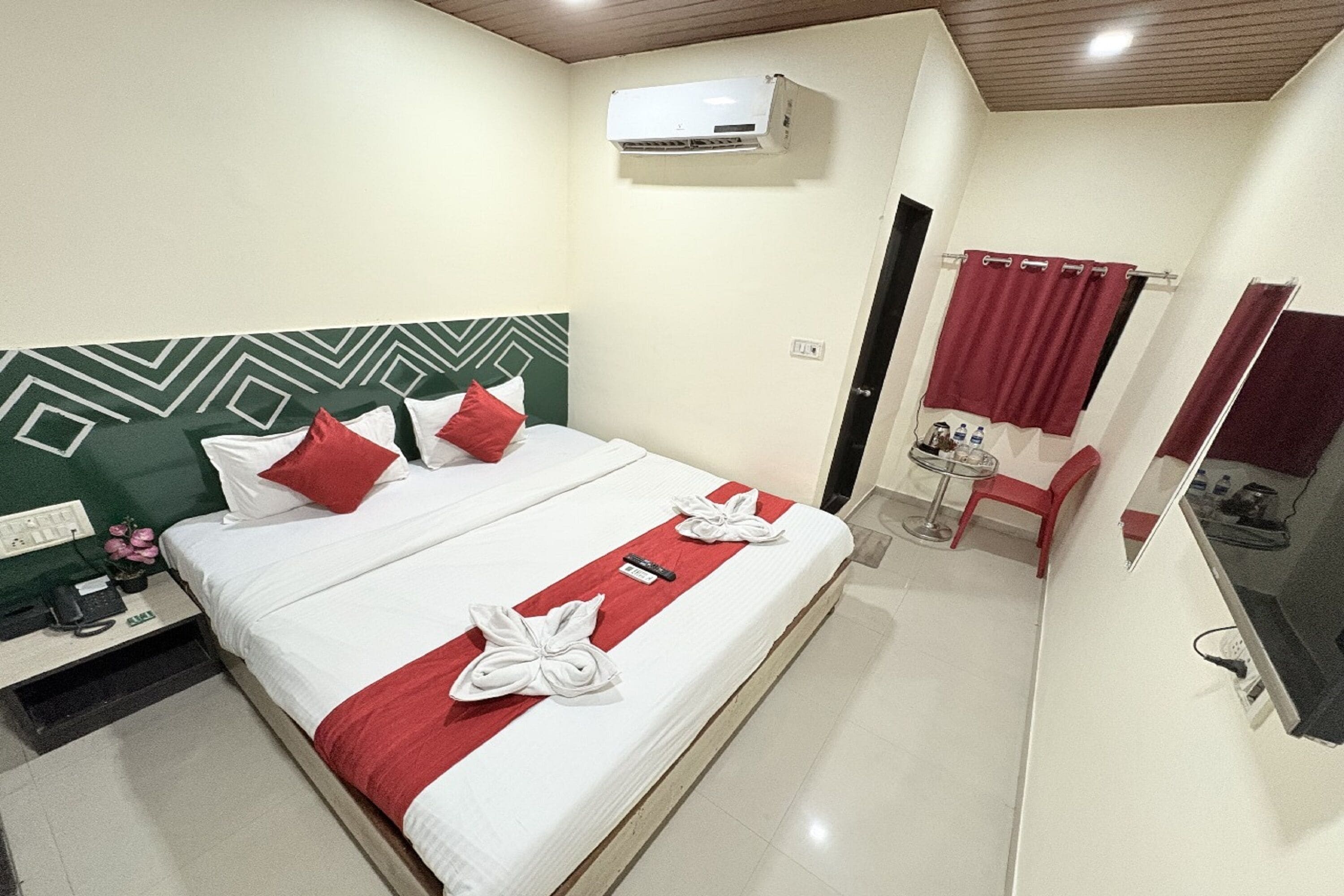 A hotel room with a bed, a desk with a phone, a chair, a small table, an air conditioning unit, and a mirror.