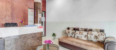 Un área de recepción con un mostrador de madera, un sofá con cojines estampados, una mesa de vidrio con un florero y un equipo de aire acondicionado en la pared.