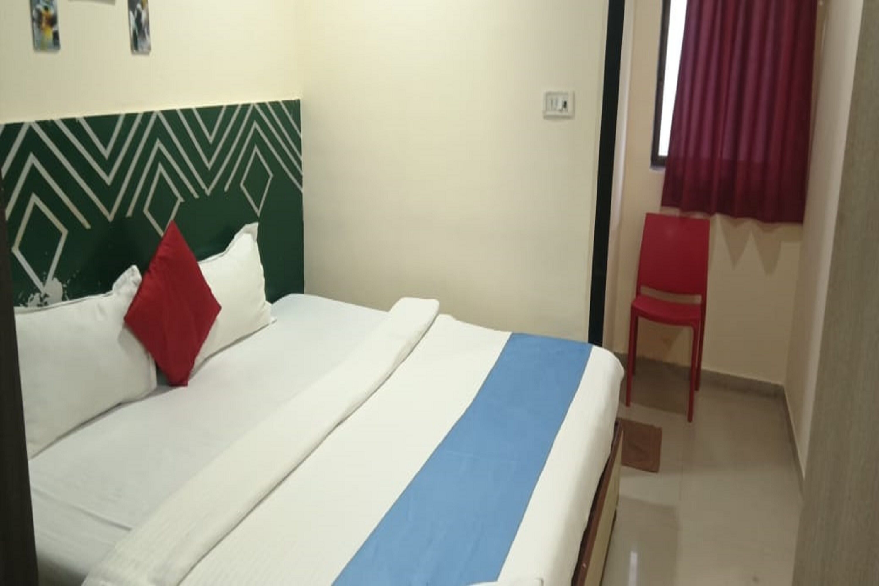 A bed with a white and blue striped bedspread, a green and white patterned headboard, and a red pillow. There is a red chair and a window with red curtains.