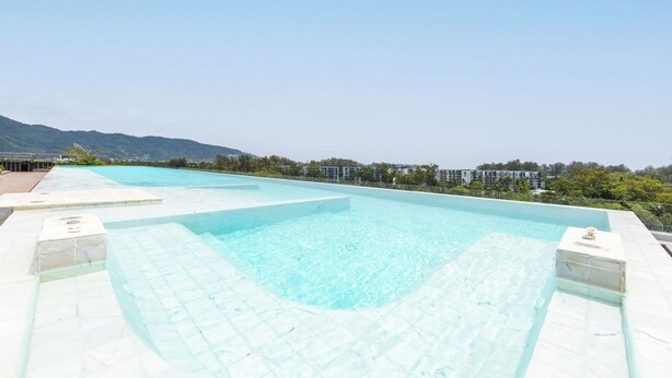A swimming pool with clear water, surrounded by a tiled deck and a view of buildings and hills in the background. - Skypark Aurora Laguna Phuket (Choeng Thale)