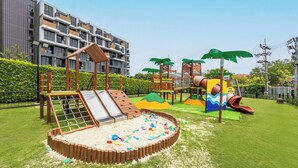A playground with slides and climbing structures, surrounded by a well-maintained lawn and a building in the background. - Skypark Aurora Laguna Phuket (Choeng Thale)