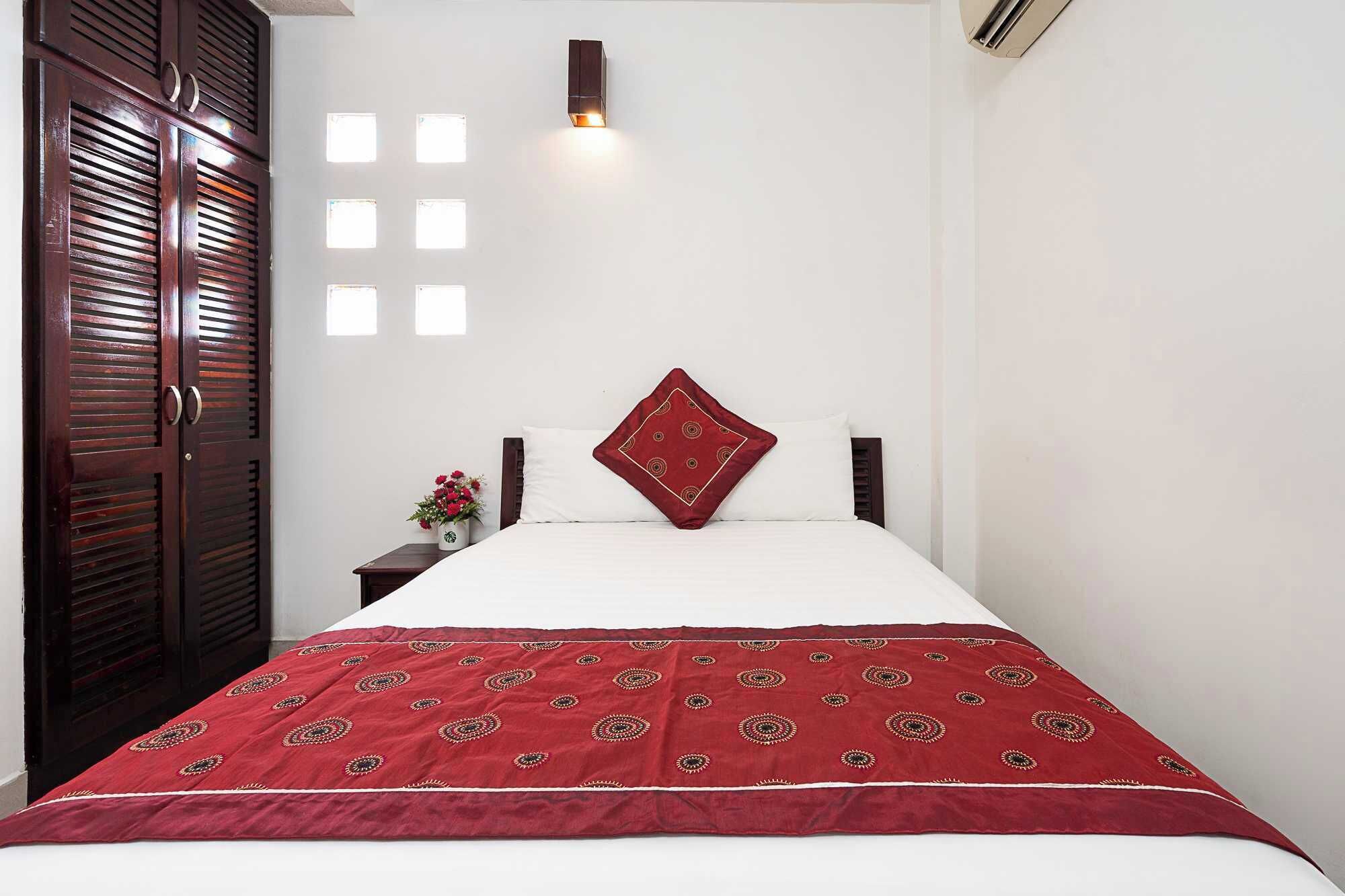 A bedroom with a bed, a red patterned bedspread, a white pillow, a red decorative pillow, a nightstand with a vase of flowers, and a wall-mounted light fixture.