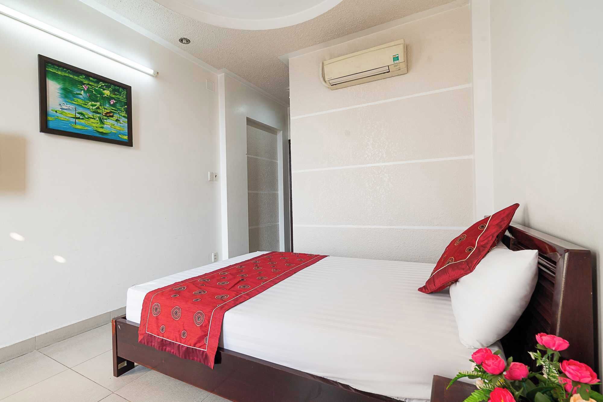 A bedroom with a bed, a red throw blanket, a pillow, a wall-mounted air conditioner, and a painting on the wall.