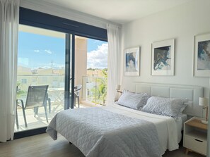 A bedroom with a bed, a nightstand, a chair, and a balcony with a view of buildings and trees.