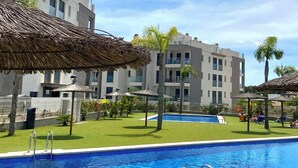 A swimming pool with a clear blue water, surrounded by a well-maintained lawn and a building with balconies in the background. - Modern 2 Bedroom Apartment in Villamartin 3 Pools, Padel, Jacuzzi Sandy Beaches (Villamartín)