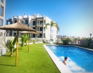 A swimming pool area with a thatched umbrella, green lawn, and modern buildings in the background.