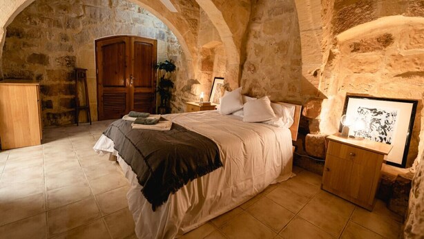 Une chambre avec une arche en pierre, un lit recouvert d’une couverture noire et blanche, une armoire en bois, une table de chevet avec une lampe et un tableau encadré accroché au mur.