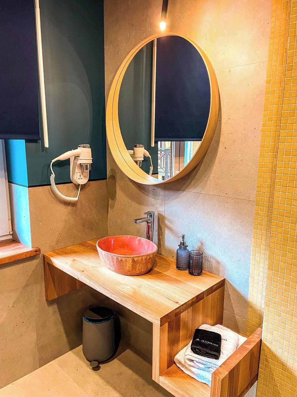 A bathroom with a wooden vanity, a round mirror, a red basin, a towel rack, and a trash bin.