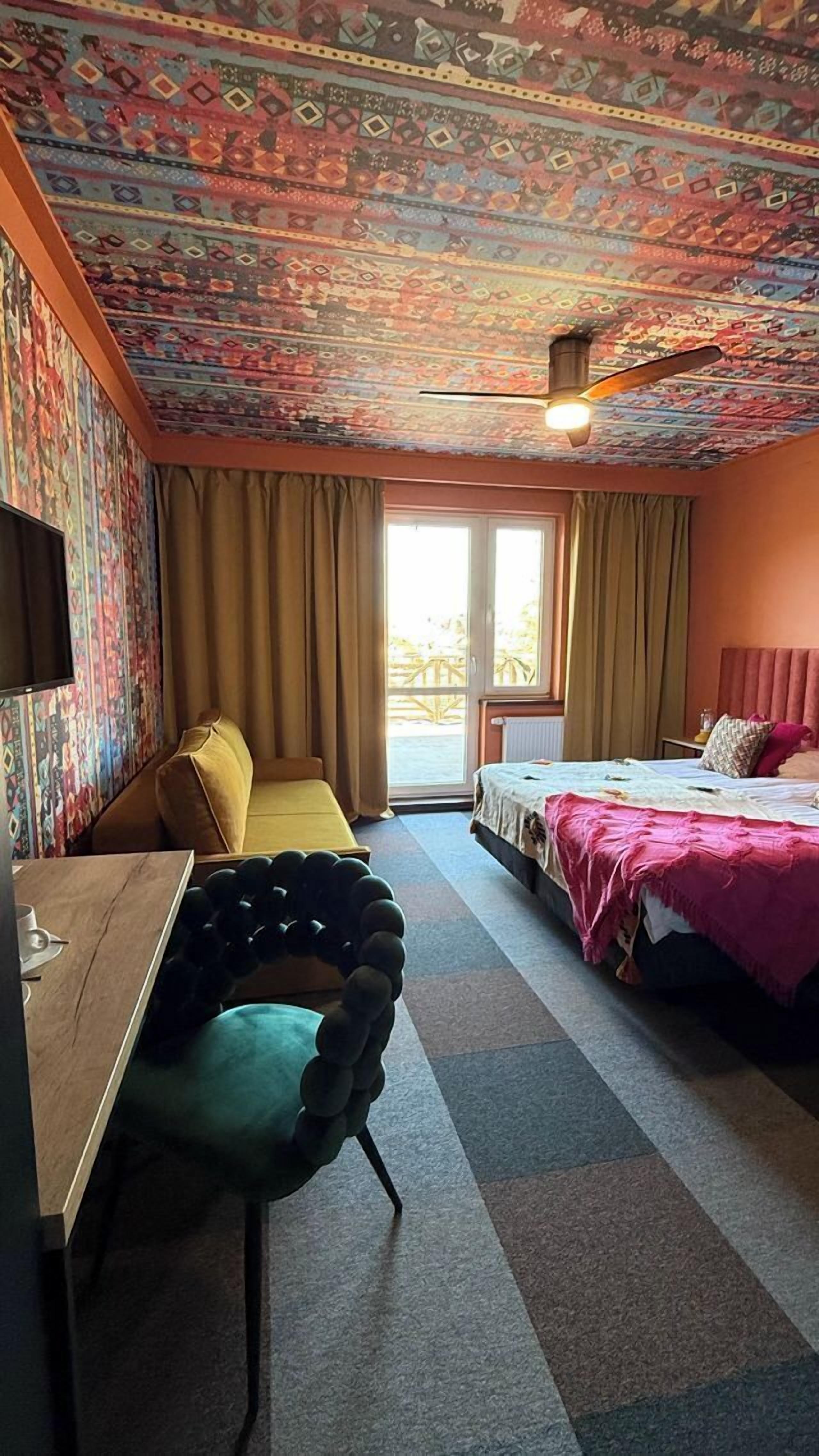 A hotel room with a patterned ceiling, a bed with a pink bedspread, a yellow chair, a desk with a lamp, and a view of the outdoors through a sliding door.