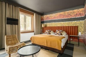 A hotel room with a bed, a round table, a wicker chair, a TV mounted on the wall, and a patterned wall. - Przystanek Tleń (Tleń)