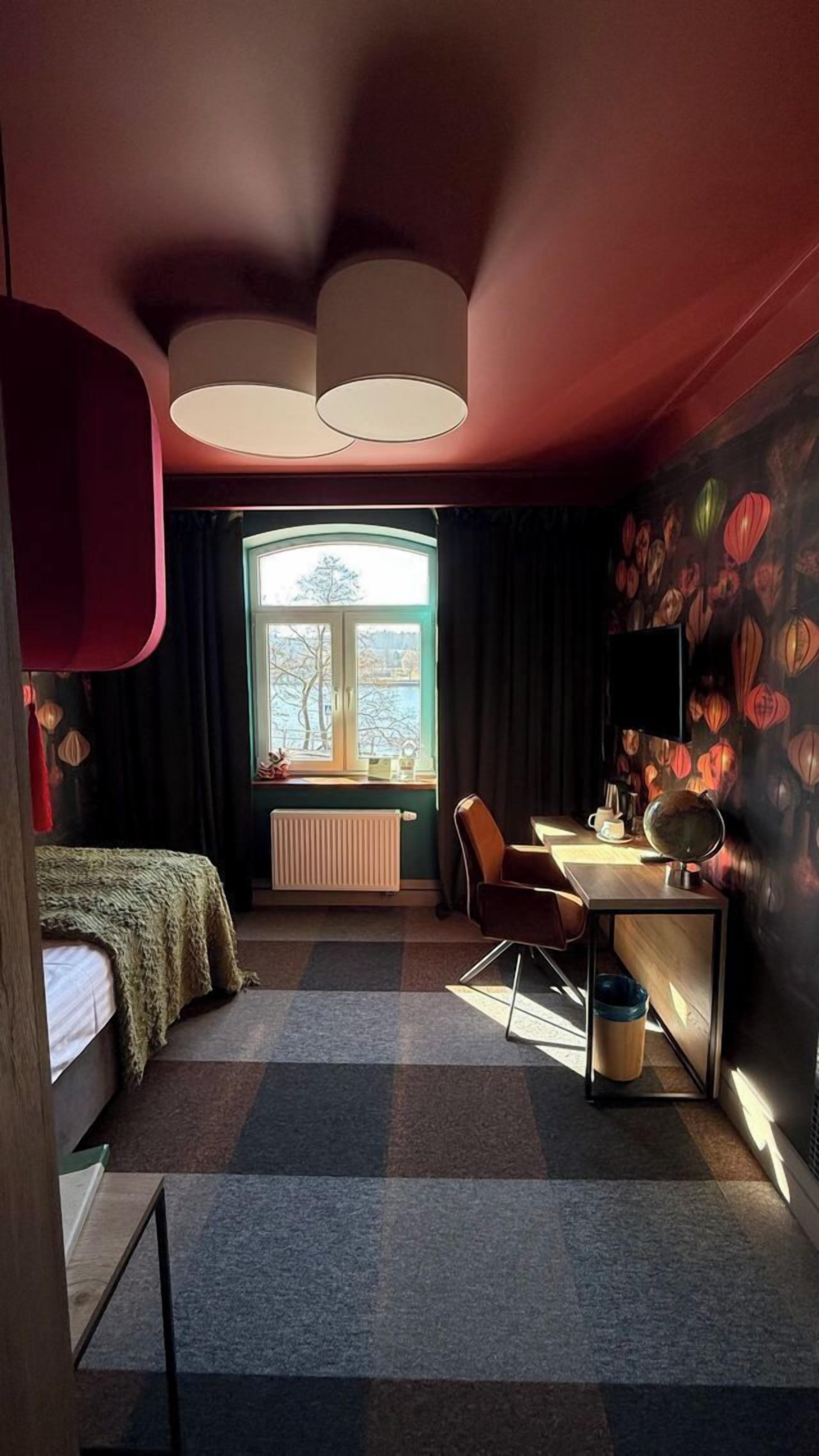 Quarto com cama, escrivaninha, cadeira, radiador e janela com vista para árvores.