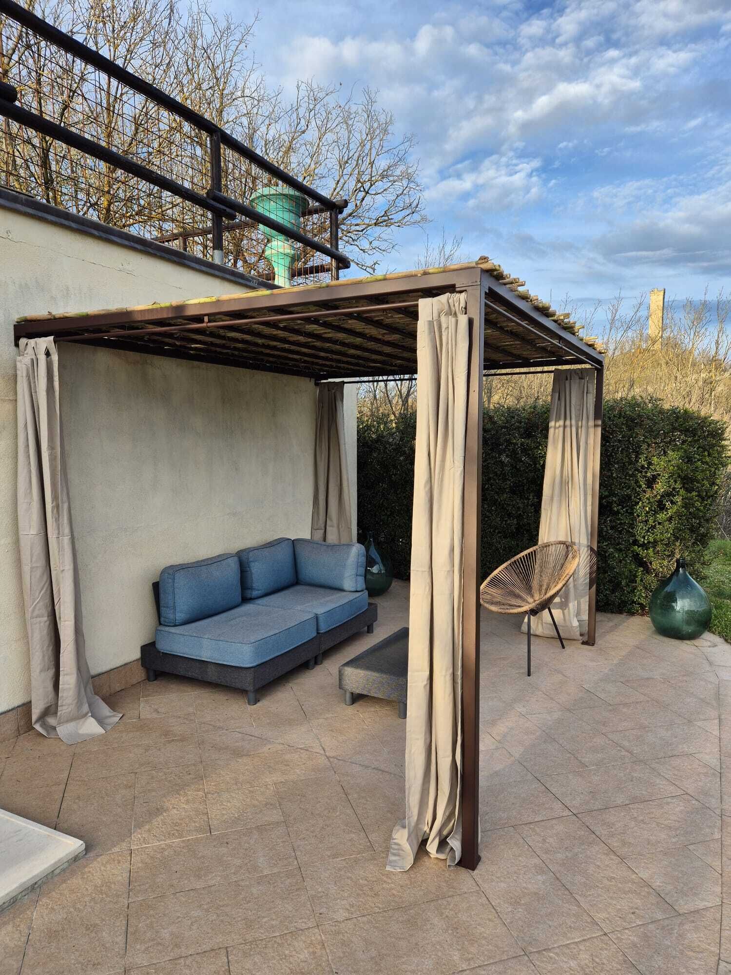 A covered patio area with a blue sofa, a wicker chair, and a large green vase.