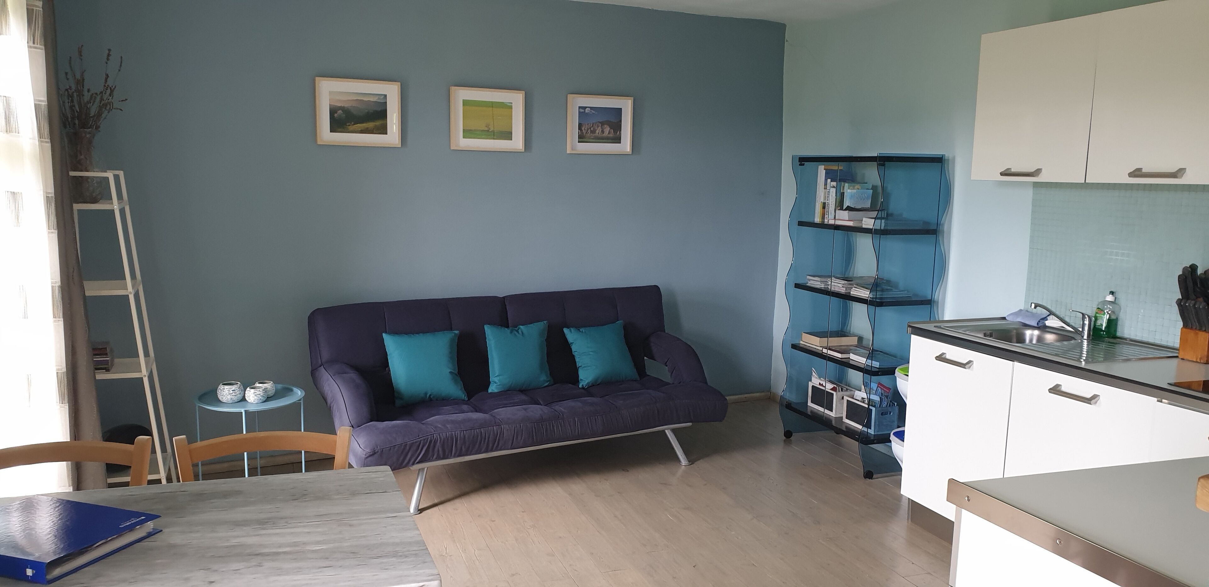 Un espacio habitable moderno con un sofá azul oscuro, una mesita redonda y una cocina con armarios blancos y encimera azul.