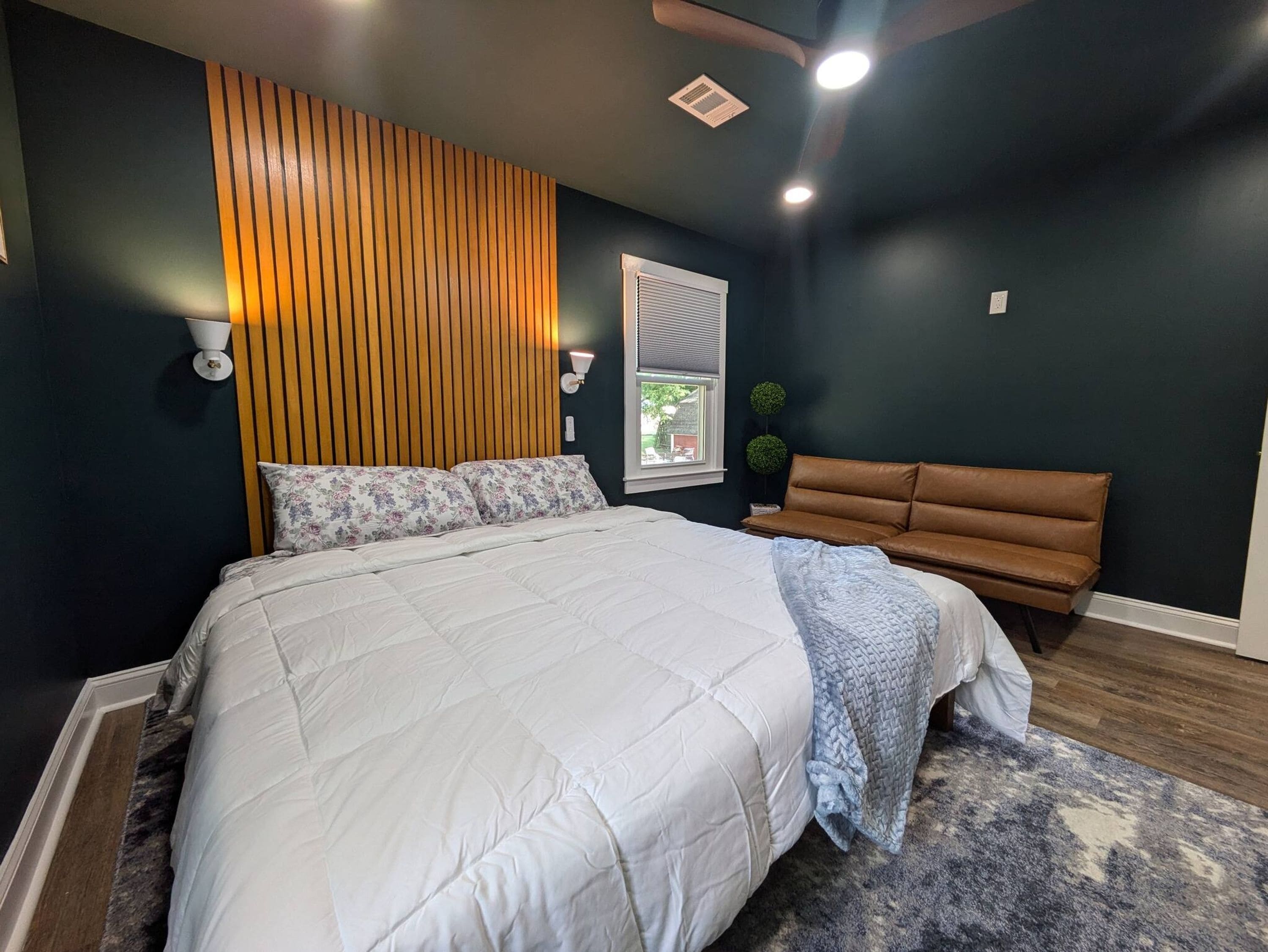Quarto com cama grande, cabeceira de ripas de madeira, um banquinho de couro marrom e janela com persianas.