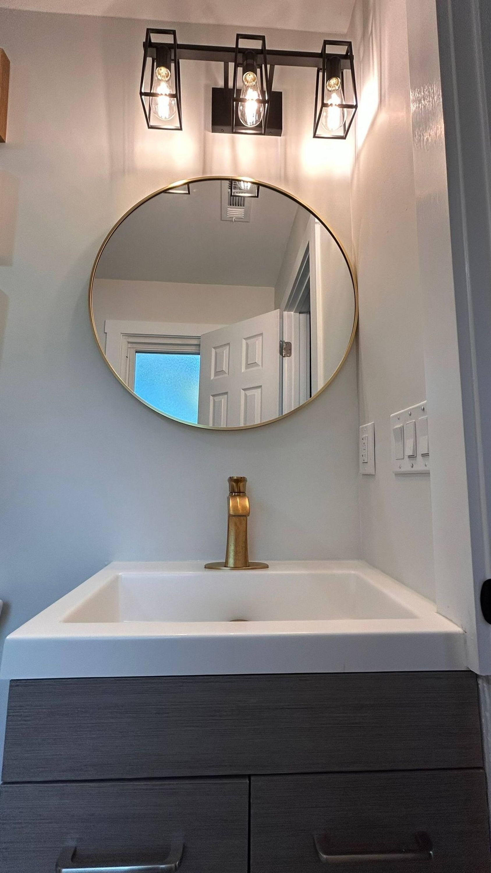 A bathroom with a white sink, a gold faucet, and a round mirror above it. There are three wall-mounted lights above the mirror.