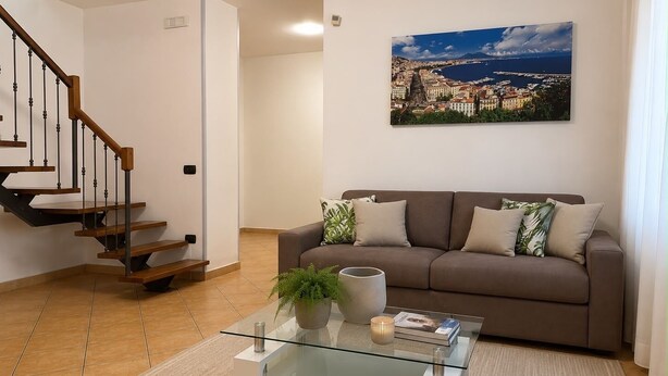 Ein Wohnzimmer mit braunem Sofa, einem Glastisch, einem Blumentopf und einem Gemälde einer Küstenstadt.