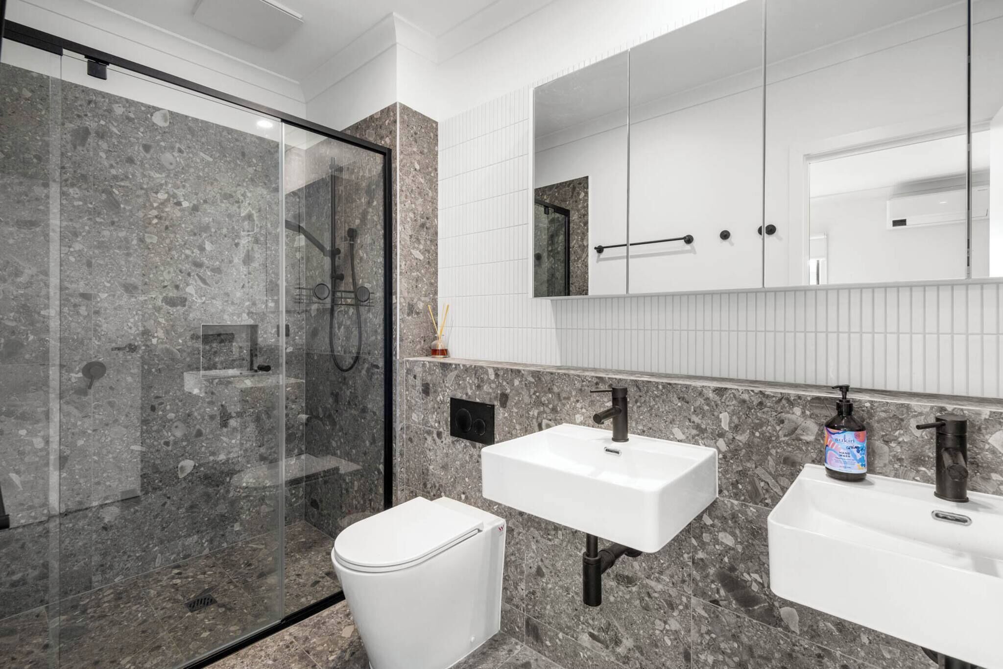 A modern bathroom with a glass-enclosed shower, a white toilet, and two sinks with a large mirror above.