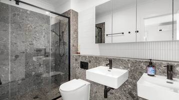 A modern bathroom with a glass-enclosed shower, a white toilet, and two sinks with a large mirror above.