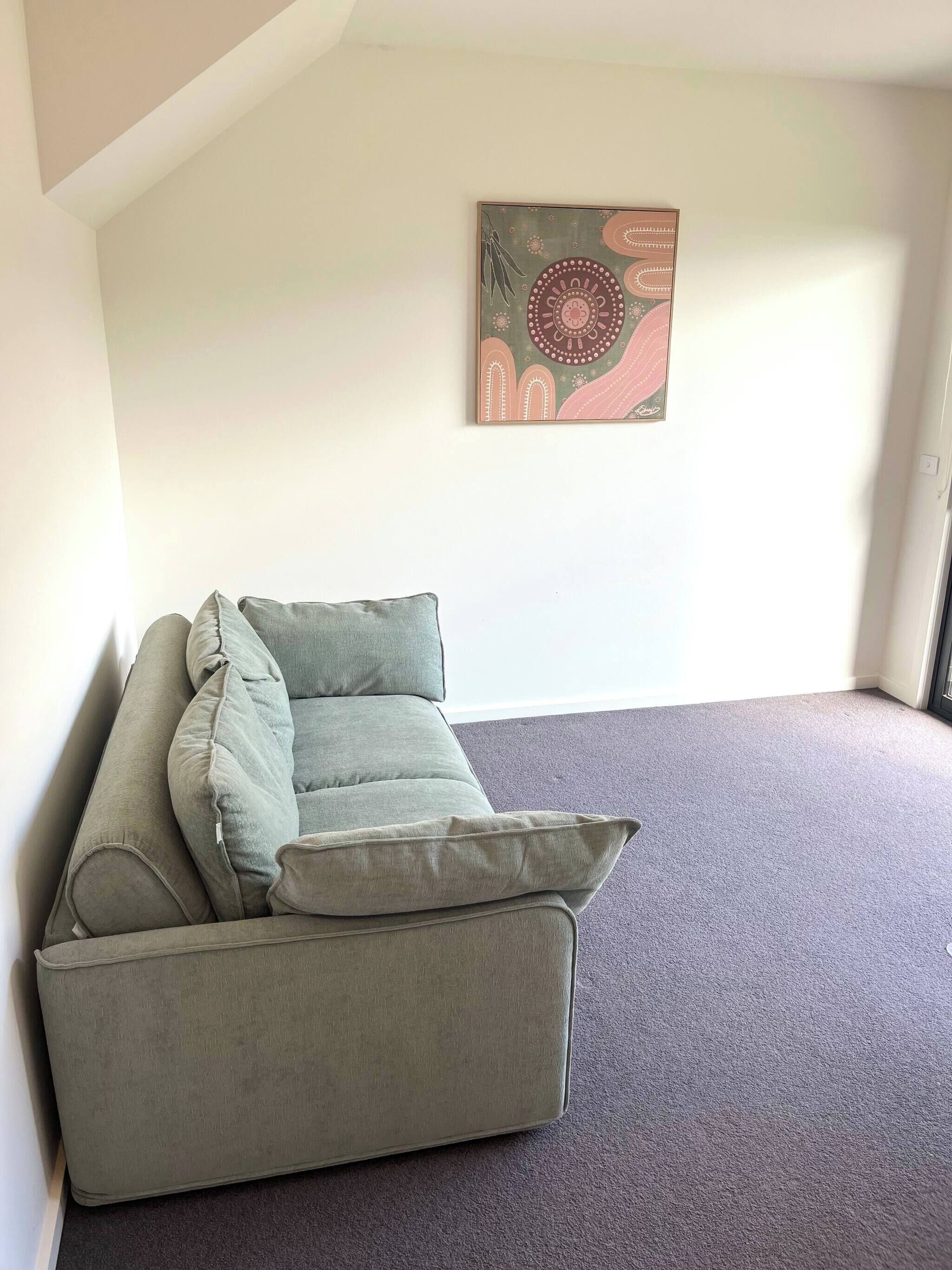 Un sofá gris con varios cojines en una habitación con piso alfombrado y una obra de arte abstracta colgada en la pared.