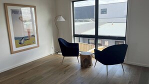 Una habitación moderna con un ventanal grande, dos sillas azul oscuro, una mesita de madera y un cuadro enmarcado en la pared.