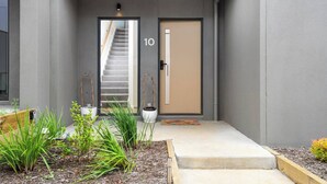 Una entrada de edificio moderno con una escalera que sube al segundo piso, una puerta con el número '10' y un pequeño jardín con plantas y un banco de madera.
