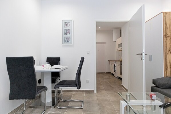 A modern, well-lit room with a dining table, two chairs, a glass coffee table, and a sofa. A kitchen area is visible in the background. - Stylish 1BR Studio In Central Vienna (Vienna)