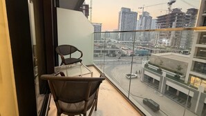 A balcony with a wicker chair and a glass table, overlooking a cityscape with buildings and construction cranes. - Azizi  405 Fully Furnished Apartment (Dubai)