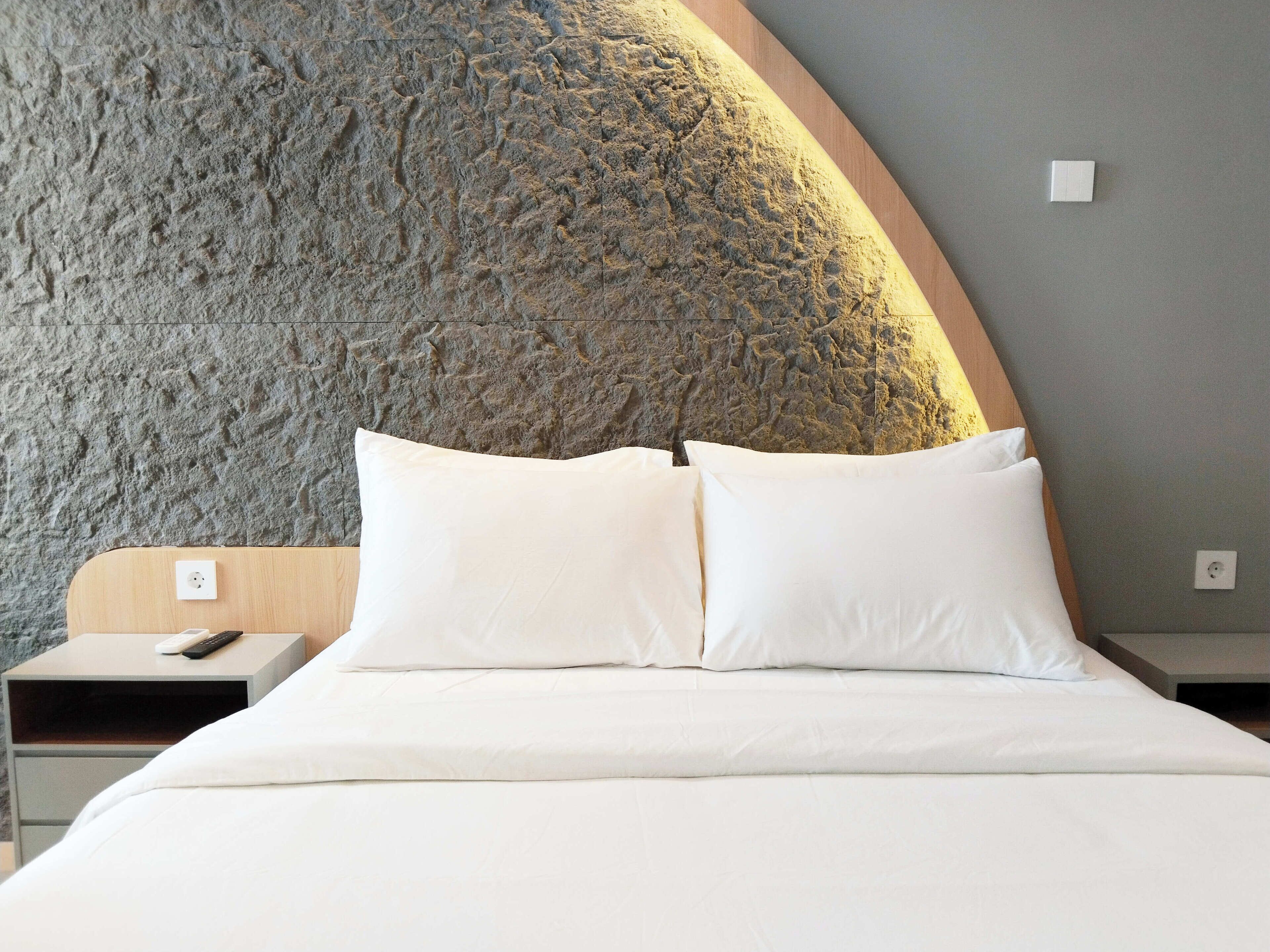 A bed with white linens, a wooden headboard, and a stone-textured wall behind it.