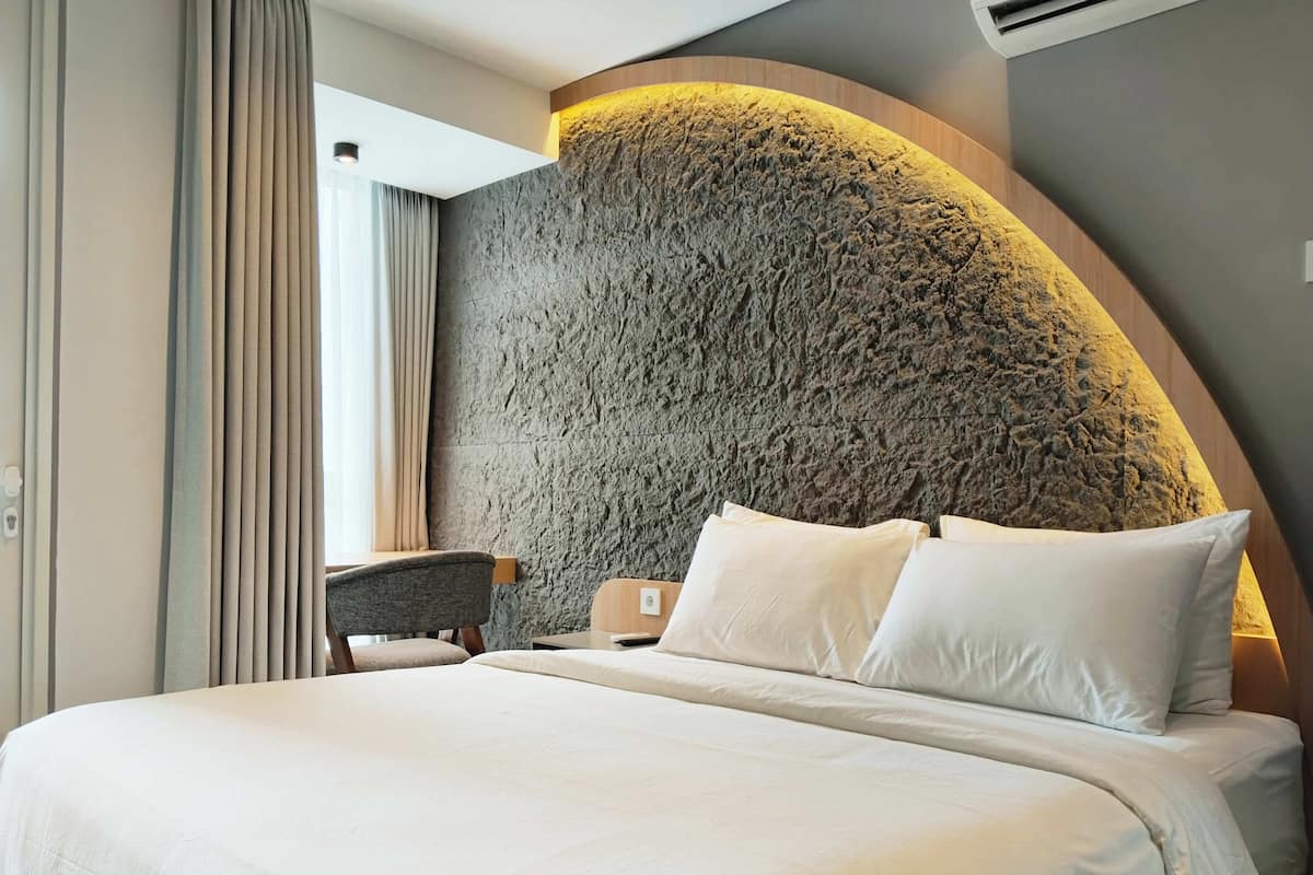 Una habitación de hotel moderna con una cama grande, una pared curva con acabado tipo piedra texturizada, un escritorio pequeño y una silla.