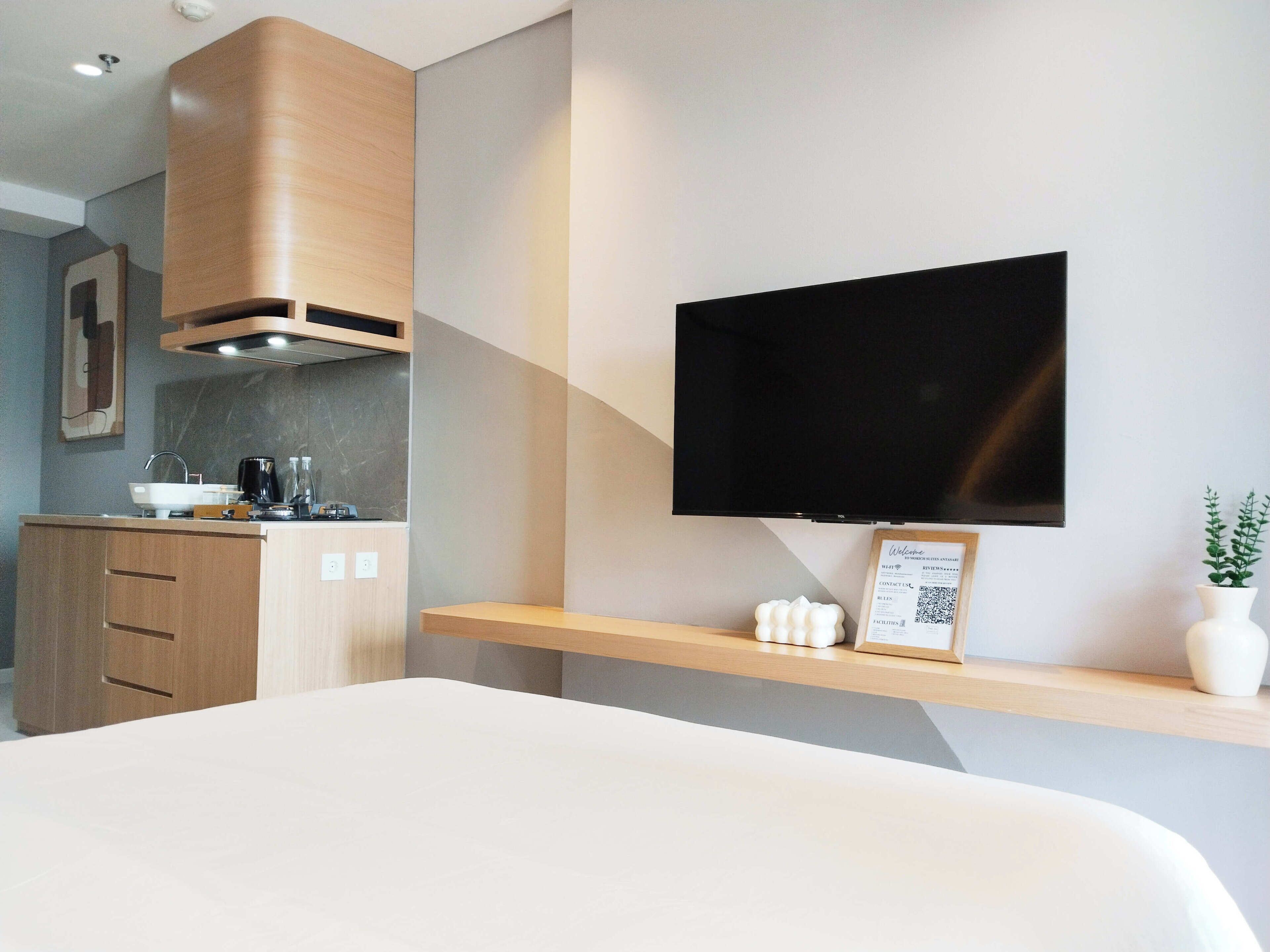 A modern hotel room with a bed, a TV mounted on the wall, a kitchen area with a sink and cabinets, and a small plant on the bedside table.