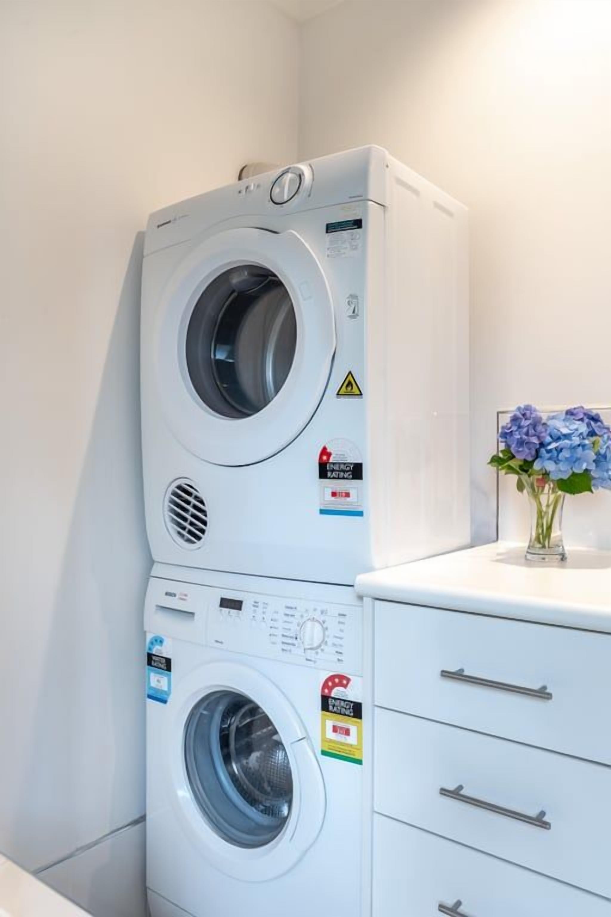A stacked washer and dryer unit with a control panel, a warning label, and a small vase with flowers on top.