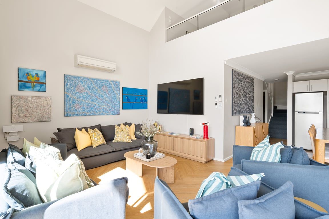A modern living room with a grey sofa, blue armchairs, and a coffee table. There is a flat-screen TV mounted on the wall, and artwork is displayed on the walls.