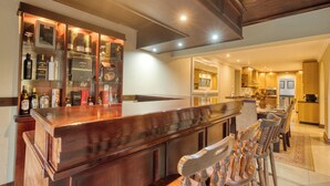 A bar area with a wooden counter and bar stools, a wine rack with bottles, and a kitchen in the background. - A spacious, private home tucked just moments from Umhlanga’s coastline. (Durban)