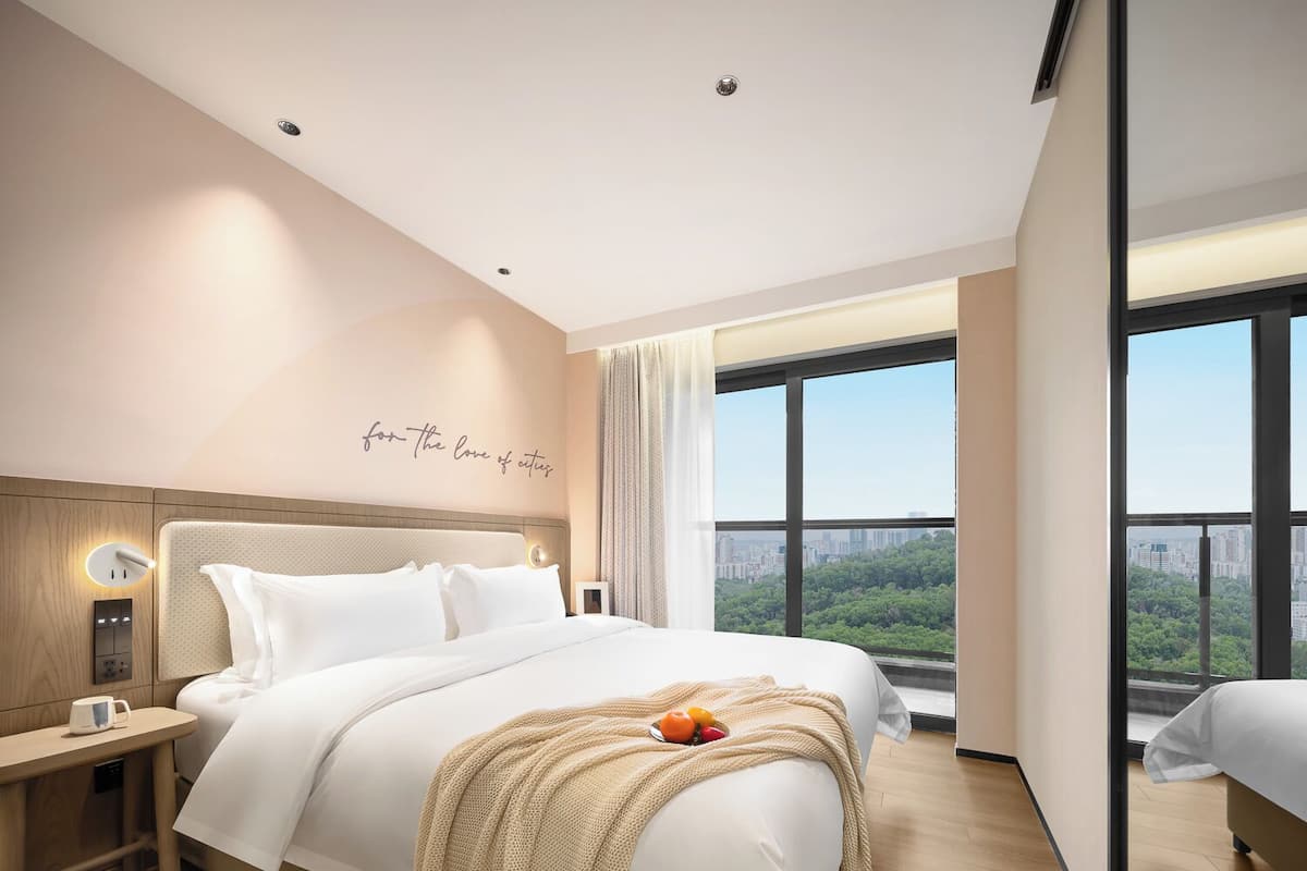 A modern hotel room with a large bed, a bedside table with a coffee cup, a wall-mounted light, and a city view through large windows.