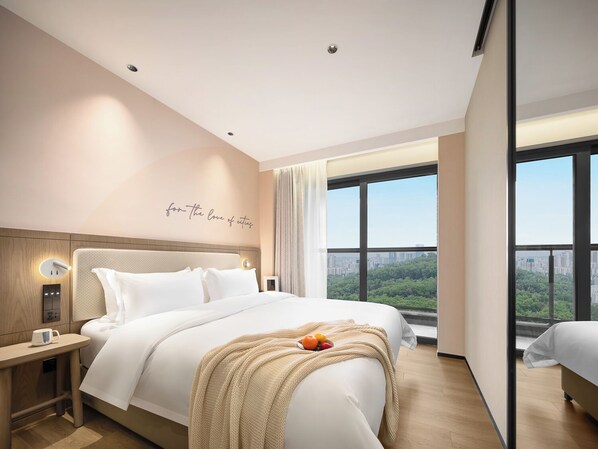 Una habitación de hotel moderna con una cama grande, una mesita de noche con una taza de café, una lámpara de pared y vistas urbanas a través de ventanas amplias.