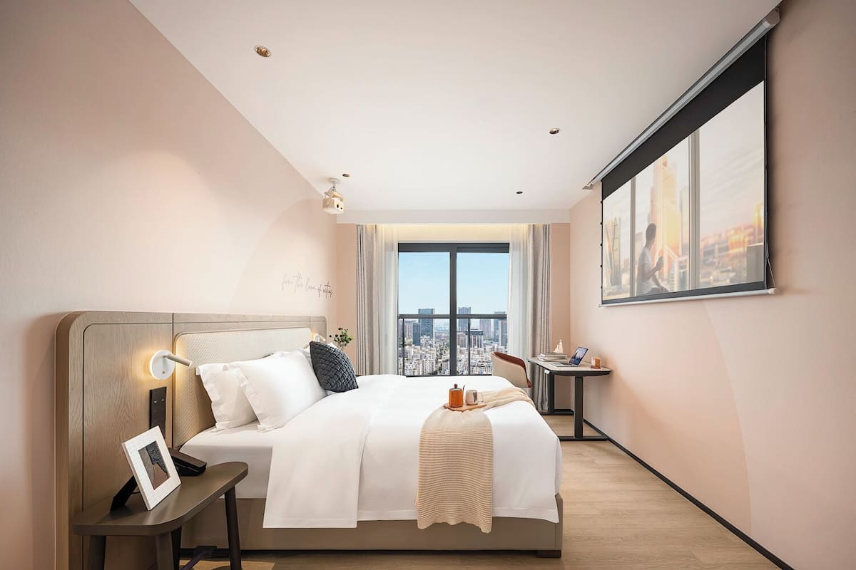 A modern hotel room with a large bed, bedside table, and a desk with a chair.